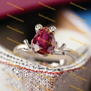 May include: A silver ring featuring a frog design. The ring has a pear-shaped, deep red gemstone as the frog's body, with two small white gemstones for eyes. The ring's band and frog details are silver. The brand name "Shehzadijewels" is visible.