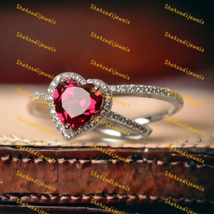 Può includere: Un anello con rubino a forma di cuore con un alone di piccoli diamanti. L'anello è in argento e ha una fascia delicata.