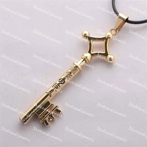 May include: A gold-colored key pendant necklace with a black cord. The key has intricate details and a decorative design. The key is about 2 inches long.