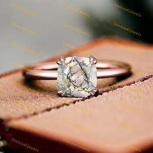May include: A rose gold ring with a square-shaped, clear gemstone with black and white veins. The ring is set on a wooden surface.