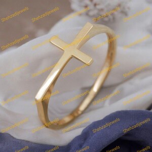 Könnte beinhalten: Ein goldfarbenes Armreif-Armband mit einem Kreuz-Design. Das Kreuz befindet sich oben auf dem Armband. Das Armband hat eine glatte, polierte Oberfläche und wird auf weißem und blauem Stoff präsentiert.