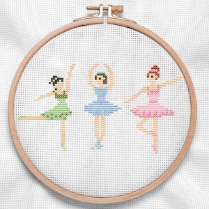 Puede incluir: Bordado de punto de cruz de tres bailarinas en un aro de madera. Las bailarinas visten tutús verdes, azules y rosas, con accesorios para el pelo a juego. El bordado está sobre tela blanca.