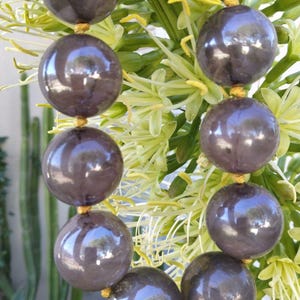 Puede incluir: Primer plano de un collar con grandes cuentas esféricas de color gris oscuro ensartadas con hilo dorado. Las cuentas tienen un acabado brillante y reflejan el entorno. El collar se encuentra sobre un fondo de flores verdes claras y amarillas.