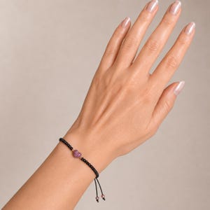 May include: Black beaded bracelet with a pink heart-shaped charm. The bracelet is on a black cord with adjustable ends. The heart charm is covered in small pink stones.