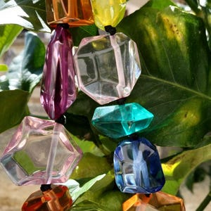 Puede incluir: Un collar colorido con cuentas geométricas facetadas en tonos naranja, amarillo, rosa, morado, verde, azul y transparente. Las cuentas están ensartadas en un cordón negro, sobre un fondo de hojas verdes.