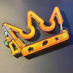 May include: An orange acrylic keychain shaped like a crown with a gun-like attachment on the left side. The base features cut-out shapes including a star and a spade. A silver metal carabiner is attached to the right side of the crown structure.