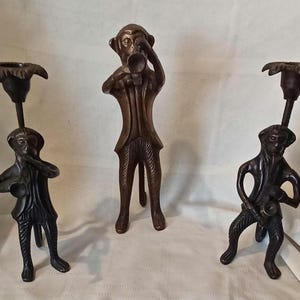 May include: Three bronze monkey candle holders. Two are playing instruments, and one is standing with a trumpet. The candle holders have a dark brown finish and are set against a white background.