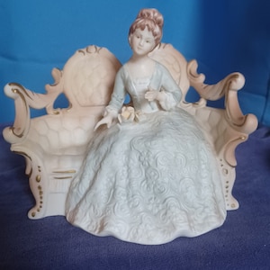 May include: A porcelain figurine of a woman in a long white dress seated on a decorative, ornate sofa. The figurine has a light blue bodice and a bun hairstyle. The sofa is light pink with gold accents.