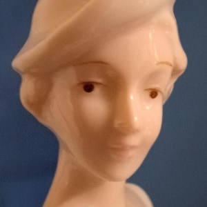 May include: A glossy porcelain figurine of a person with a smooth, pale complexion, dark eyes, and a soft expression. The figure wears a sculpted hat with a wide, curved brim that rests atop the head, framing the face with elegant, simple lines.