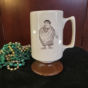 May include: Cream-colored ceramic mug with a brown pedestal base and a large handle. The mug features a black and white illustration of a Roman figure. The mug is likely used for drinking beverages.
