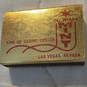 May include: A rectangular gold-colored matchbox featuring red text that reads DEL WEBB'S MINT, KING OF CASINO CENTER, and LAS VEGAS, NEVADA. The design includes a red star graphic above the text and a red line accenting the lettering on the front.