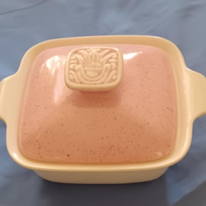 May include: A square, lidded casserole dish with a pink speckled top and a white base. The lid has a decorative, raised handle. The dish has two side handles and is set against a blue background.