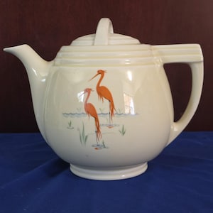 May include: A cream-colored ceramic teapot featuring a decorative illustration of two orange long-legged birds standing in blue water with green reeds. The teapot has a stepped lid, a curved spout, and a geometric handle with horizontal ridges.
