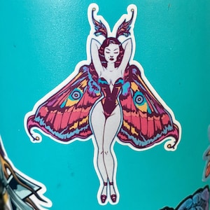 May include: A sticker featuring a stylized moth-woman design. The woman has pale skin, red hair, and is wearing a dark purple bodysuit. Her arms are raised above her head. She has large, colorful wings with pink, blue, and yellow patterns. The sticker is on a light blue surface.