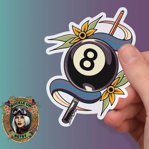 May include: A sticker of a black and white eight ball with a blue ribbon and yellow flowers. The eight ball is surrounded by a white outline. The sticker has a red and white stripe on the bottom. The text 'buckle up betsy' is visible in the background.