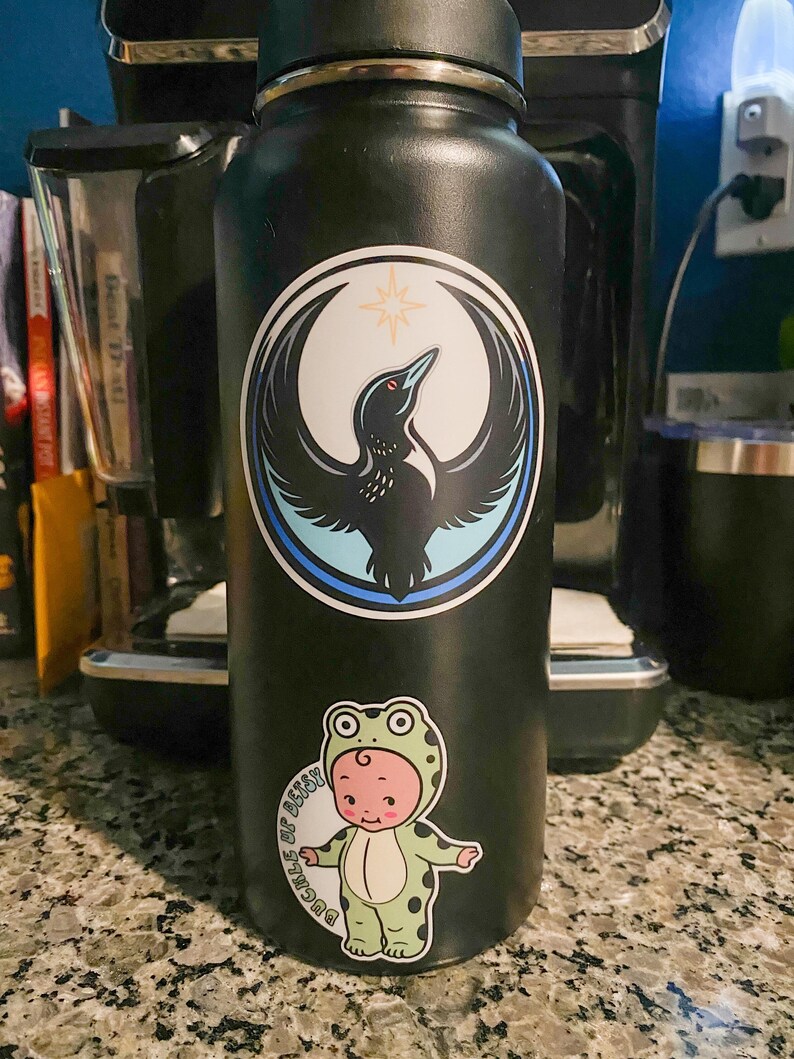 May include: A black water bottle with two stickers. One sticker features a bird with outstretched wings inside a circle. The other sticker shows a baby in a frog costume with the text "Buckles of Betsy".