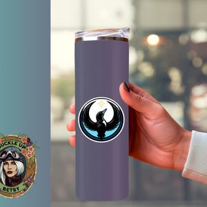 May include: A purple insulated tumbler with a black and white bird emblem. The emblem is a circular design with a bird silhouette, a crescent moon, and a star. The tumbler is held by a person.