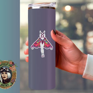Peut inclure: Un gobelet isotherme violet avec un autocollant de papillon color&eacute;. Le motif de papillon pr&eacute;sente le corps d'une femme avec des ailes de papillon dans des tons de rose, jaune et violet. Le gobelet est tenu par une personne.