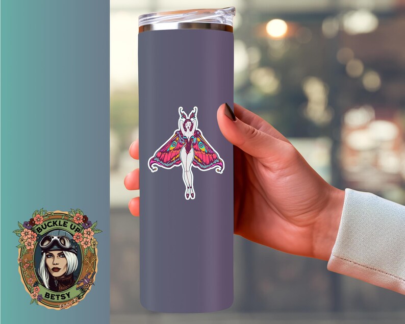 Peut inclure: Un gobelet isotherme violet avec un autocollant de papillon color&eacute;. Le motif de papillon pr&eacute;sente le corps d'une femme avec des ailes de papillon dans des tons de rose, jaune et violet. Le gobelet est tenu par une personne.