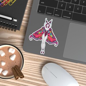 Peut inclure: Un autocollant color&eacute; repr&eacute;sentant une femme avec des ailes de papillon, dans des tons de rose, jaune et bleu, est coll&eacute; sur un ordinateur portable. L'image est pos&eacute;e sur un bureau en bois, &agrave; c&ocirc;t&eacute; d'une tasse de chocolat chaud avec des guimauves et des b&acirc;tons de cannelle.