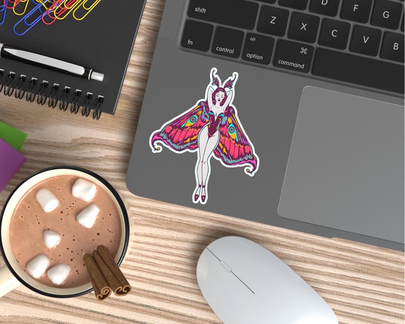 Peut inclure: Un autocollant color&eacute; repr&eacute;sentant une femme avec des ailes de papillon, dans des tons de rose, jaune et bleu, est coll&eacute; sur un ordinateur portable. L'image est pos&eacute;e sur un bureau en bois, &agrave; c&ocirc;t&eacute; d'une tasse de chocolat chaud avec des guimauves et des b&acirc;tons de cannelle.