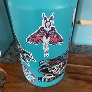 Peut inclure: Une gourde turquoise orn&eacute;e de plusieurs autocollants. L'autocollant central repr&eacute;sente une femme avec des ailes de papillon, dans un sch&eacute;ma de couleurs rouge et violet. D'autres autocollants incluent un cr&acirc;ne et un papillon de nuit.