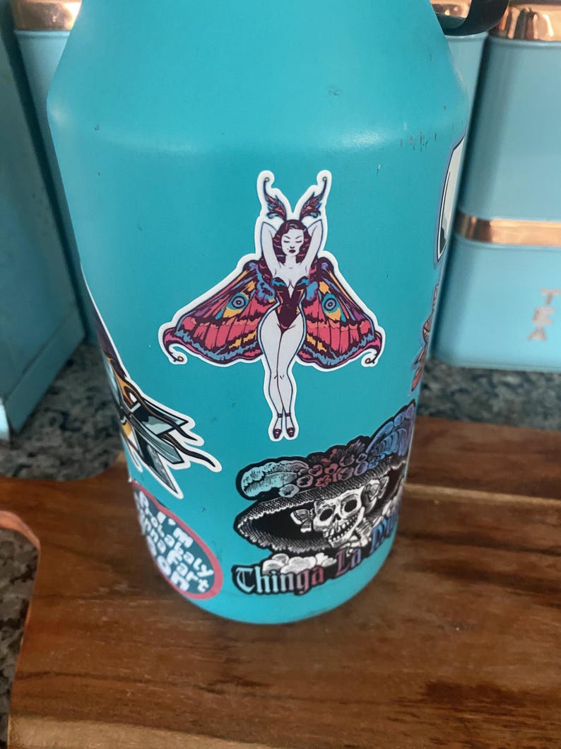Peut inclure: Une gourde turquoise orn&eacute;e de plusieurs autocollants. L'autocollant central repr&eacute;sente une femme avec des ailes de papillon, dans un sch&eacute;ma de couleurs rouge et violet. D'autres autocollants incluent un cr&acirc;ne et un papillon de nuit.