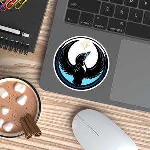 May include: A round sticker featuring a black bird with outstretched wings, a blue and white border, and a gold star. The sticker is on a laptop, with a cup of hot chocolate and a computer mouse nearby.