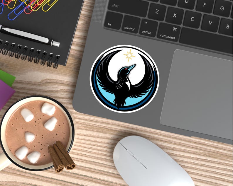May include: A round sticker featuring a black bird with outstretched wings, a blue and white border, and a gold star. The sticker is on a laptop, with a cup of hot chocolate and a computer mouse nearby.