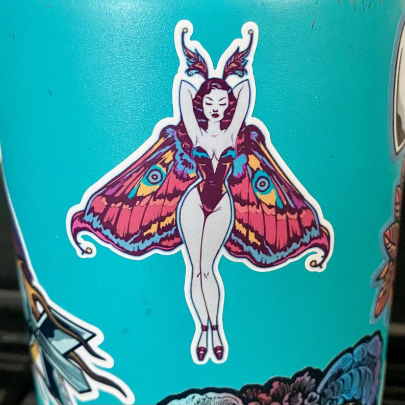 May include: A sticker featuring a stylized moth-woman design. The woman has pale skin, red hair, and is wearing a dark purple bodysuit. Her arms are raised above her head. She has large, colorful wings with pink, blue, and yellow patterns. The sticker is on a light blue surface.