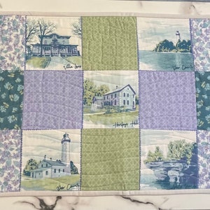 May include: A patchwork quilt table runner with a light blue, green, and purple color scheme. The runner features a variety of images, including houses, lighthouses, and floral patterns. The text "Wright's Brewers Villa Linda" is visible on the runner.