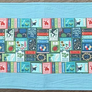 May include: A blue quilted table runner with a colorful pattern of books and flowers. The pattern is repeated across the runner.
