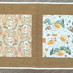 May include: A brown table runner with four square panels. Each panel features a different white and brown patterned design. The panels are separated by a brown border.
