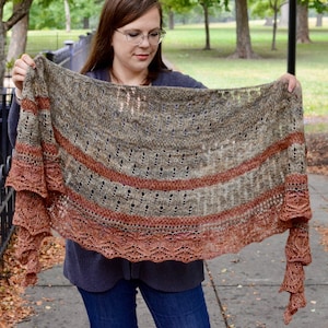 以下が含まれることがあります： レースの縁取りが施された、茶色、灰色、オレンジ色のニットのショール。ショールは、公園に立っている人の腕にかけられています。