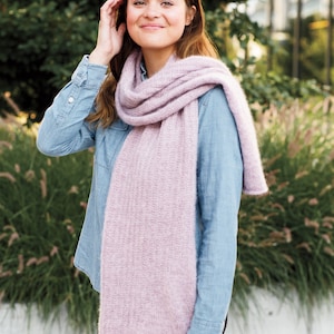 May include: A woman wearing a denim shirt and a long, light pink scarf. The scarf is knitted and has a subtle texture. She is standing in front of a green bush.