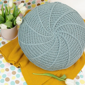 May include: A light blue crocheted round cushion with a swirl pattern. The cushion is on a yellow knitted blanket and a white background with colorful polka dots.
