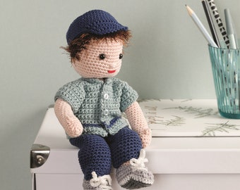 May include: A crocheted doll wearing a blue hat, blue pants, and a light blue shirt with buttons. The doll is sitting on a white box with a silver latch.