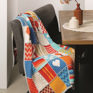 Puede incluir: Una manta de crochet colorida con un diseño de patchwork que presenta varias texturas y patrones, incluyendo corazones, árboles y formas geométricas. La manta está drapeada sobre una silla gris y una mesa de madera con un jarrón de flores secas.