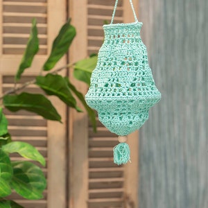 Op de afbeelding: Een lichtblauwe gehaakte hanglamp met een houten kraal en een kwast. De lamp hangt aan een witte plafondhaak.