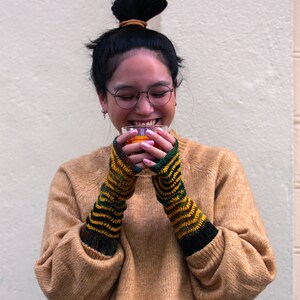 May include: A person wearing a brown sweater and black fingerless gloves is holding a glass of orange liquid. They are smiling and looking down at the glass.