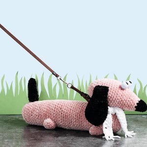 Op de afbeelding: Een roze gehaakt dackel speelgoed met zwarte oren en een witte sjaal is aan de lijn en loopt op een grijs oppervlak voor een groene gras achtergrond.