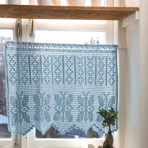 May include: A light blue crocheted window valance hangs in a window with a white wooden frame.  The valance has a scalloped bottom edge.  There are potted plants on a wooden shelf above the window.