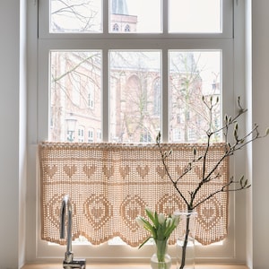 Op de afbeelding: Een witte keukengoten met een chromen kraan en een gehaakte gordijnband die in het raam hangt. Een vaas met witte tulpen en een hoge glazen vaas met een takje twijgen staan op het aanrecht.