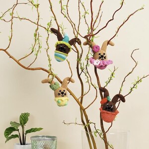 Pode incluir: Enfeites de coelhinhos da Páscoa de crochê em várias cores, pendurados em um galho de árvore. Os coelhinhos têm corpos marrons, bege e laranja com detalhes coloridos. Os galhos da árvore estão nus com pequenas folhas verdes.