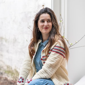 May include: Cream-colored crochet cardigan with red and blue stripes on the sleeves and floral accents on the cuffs. The cardigan has red buttons and is worn over a denim shirt and blue dress. The overall style is bohemian.