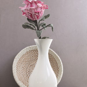 Puede incluir: Una flor de protea de crochet rosa y blanco en un jarrón de cerámica blanco. El jarrón está sobre un posavasos de mimbre tejido.