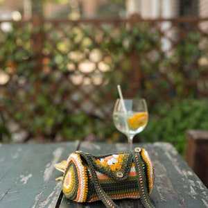 May include: A handmade crochet barrel bag in shades of green, orange, white, and yellow. The bag features a geometric pattern and a long strap. The bag is resting on a weathered wooden table. A glass of water with a straw and orange slice is in the background.