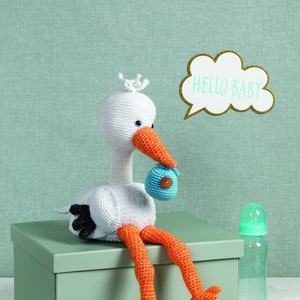 Puede incluir: Una cigüeña blanca de crochet sosteniendo un bebé azul de crochet en su pico, sentada en una caja verde con las palabras "HELLO BABY" en un globo de diálogo blanco encima. Hay tres pequeños juguetes de bebé de crochet en el suelo frente a la cigüeña.