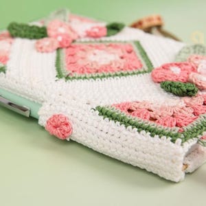 May include: A handmade white crochet case with pink and green floral accents. The case features a square pattern with a green border and a light green pen inserted. The case is designed to hold a pen or pencil.