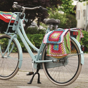Op de afbeelding: Lichtblauwe fiets met een zwarte mand en een kleurrijke gehaakte mandvoering. De fiets heeft een bijpassende gehaakte fietstas op de achterdrager. De banden hebben witte zijwanden.